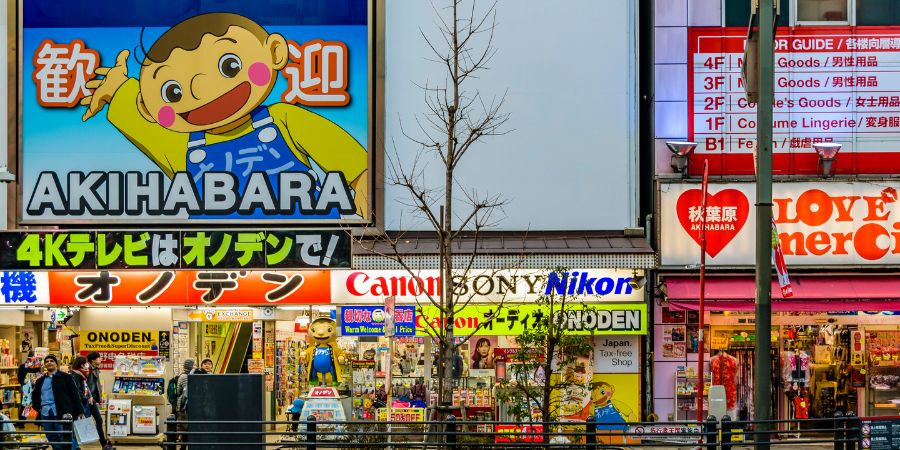 A colorful street view of the Akihabara district in Tokyo showcasing storefronts with bright signs for cameras electronics and tax free shopping. This area is a world famous hub for manga otaku and fans of Japanese pop culture who visit the many specialty bookshops and fan forums in the city.