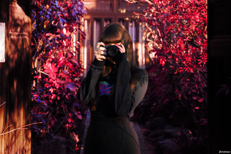 Woman stood in front of leafy building holding a camera up to her face and taking a photo.