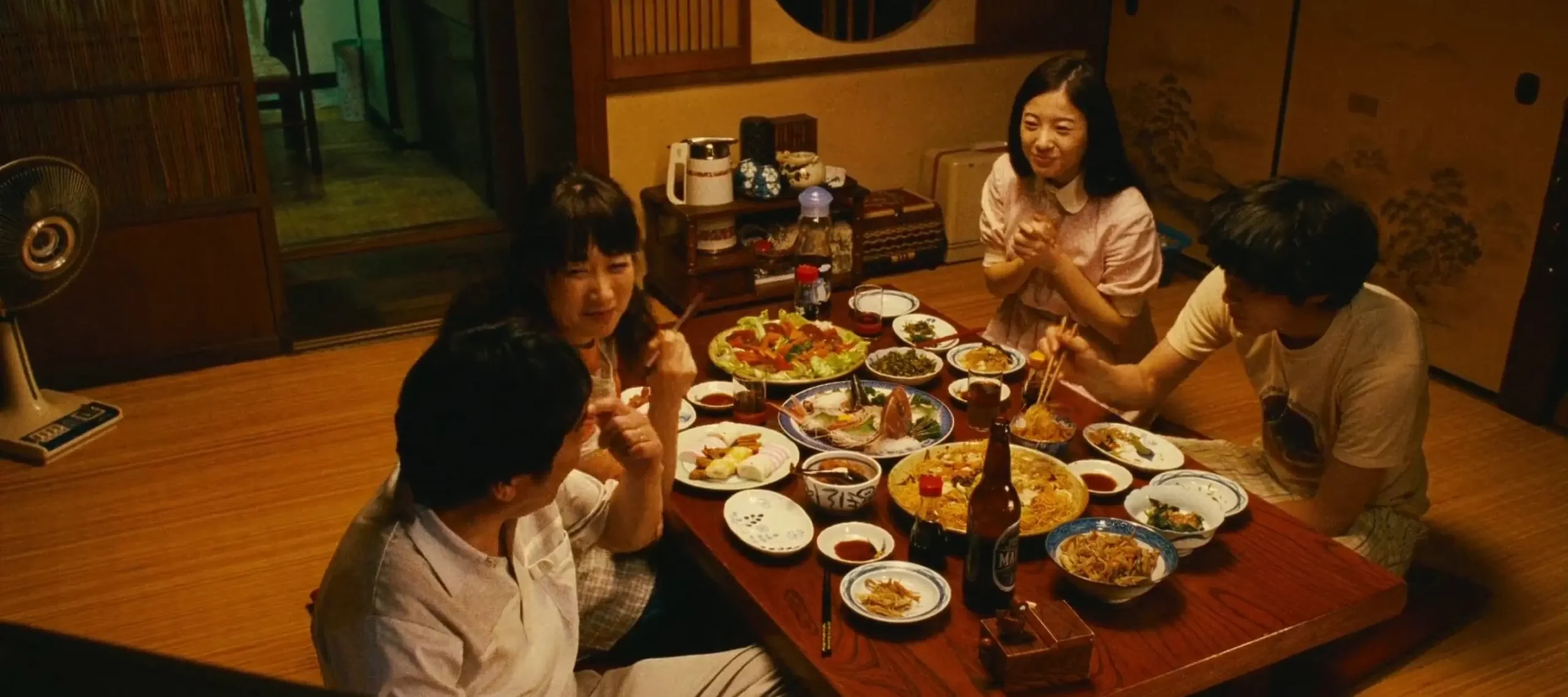 Family sharing a meal around a low table in a traditional Japanese home, with multiple dishes and drinks spread out as they chat and eat together