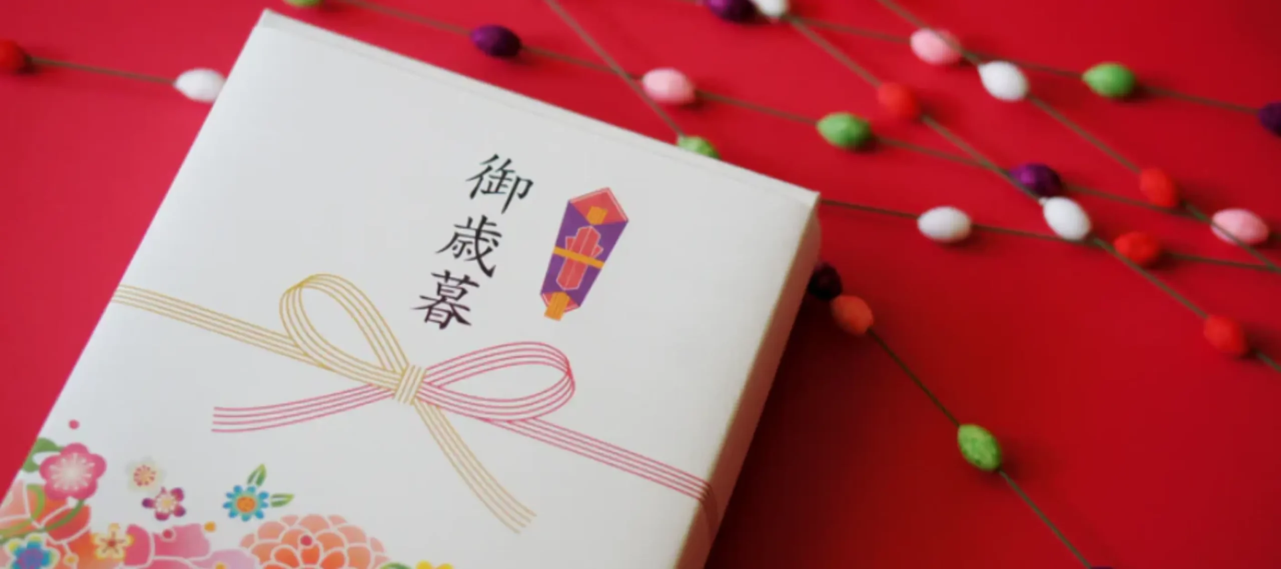 Oseibo (御歳暮) gift box wrapped with decorative mizuhiki cord, set against a red background with colourful festive ornaments