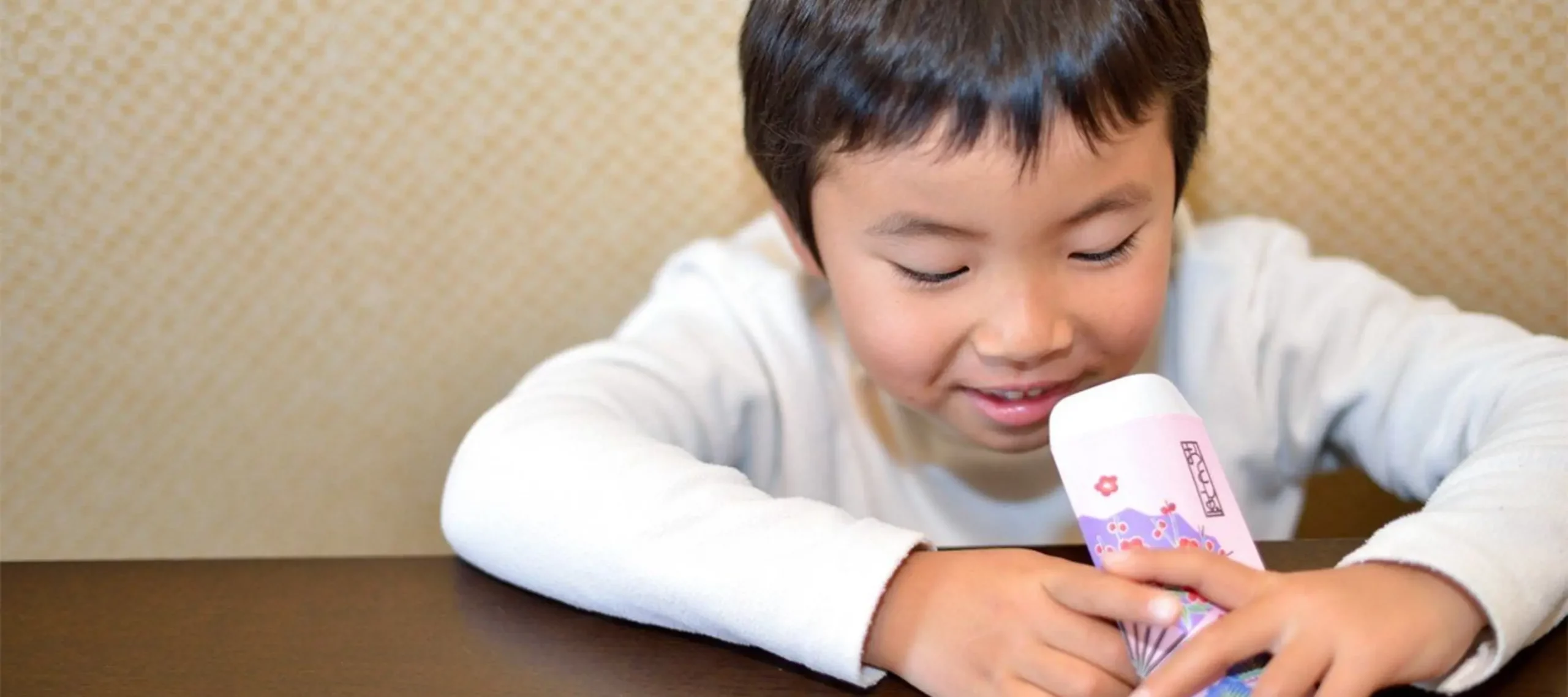 Young child smiling while holding a decorated otoshidama envelope, sitting at a table and looking down at the gift money inside