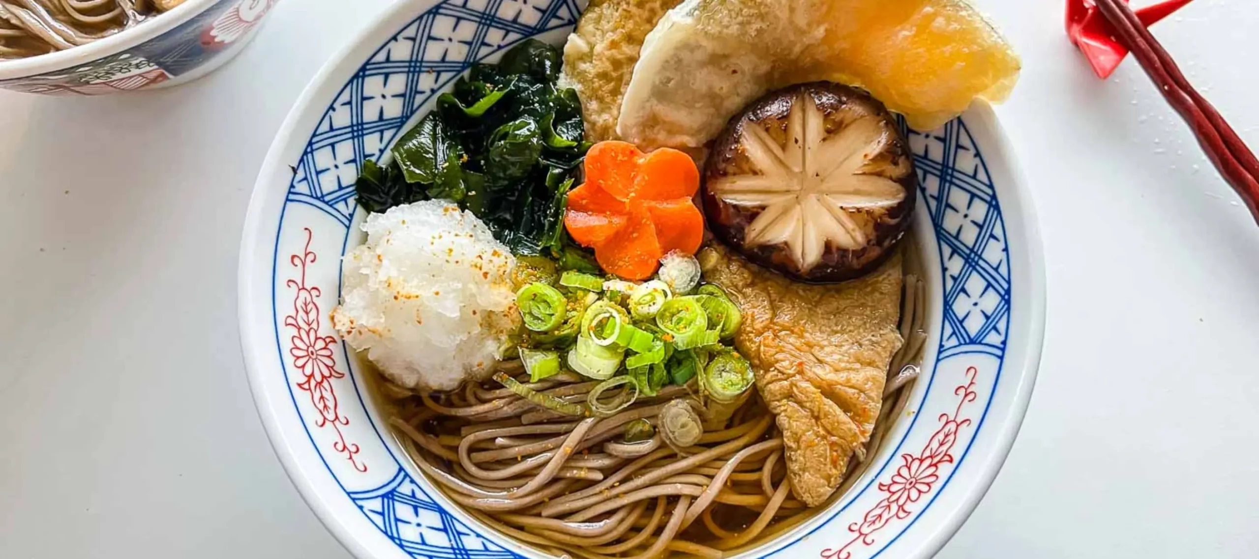 Bowl of soba noodles in broth topped with tempura, shiitake mushroom, seaweed, grated daikon, and sliced spring onions