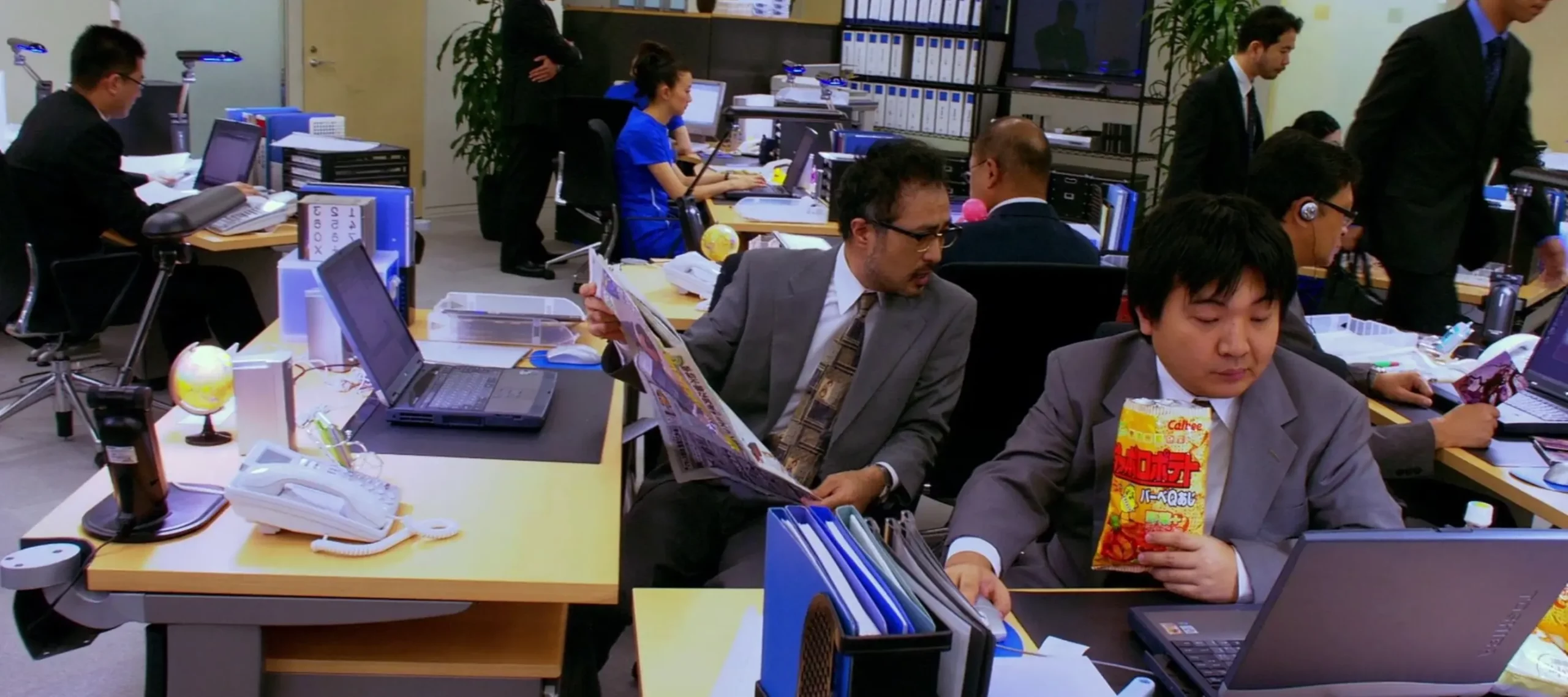 Busy Japanese office with employees at desks using laptops, one reading a newspaper while another eats snacks, surrounded by files and office equipment