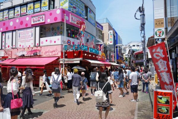Shoppers crowding the entrance to Takeshita Street in Harajuku, lined with pink shop fronts and banners on a sunny day