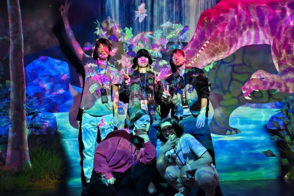 A tour group posing in front of a glowing projection of a waterfall and a giant pink dinosaur during the MagicalTrip Harajuku tour