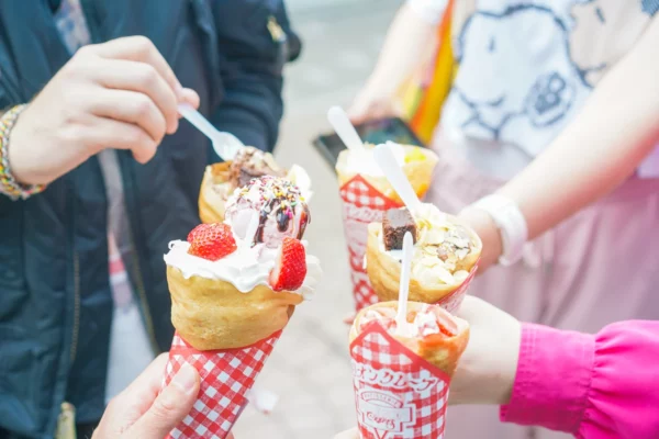 Hands holding Harajuku crepes filled with whipped cream, strawberries and ice cream in red and white checked wrappers
