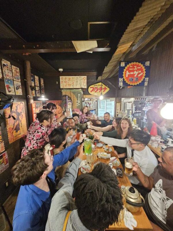 Large group raising glasses along a long table in a busy Tokyo izakaya