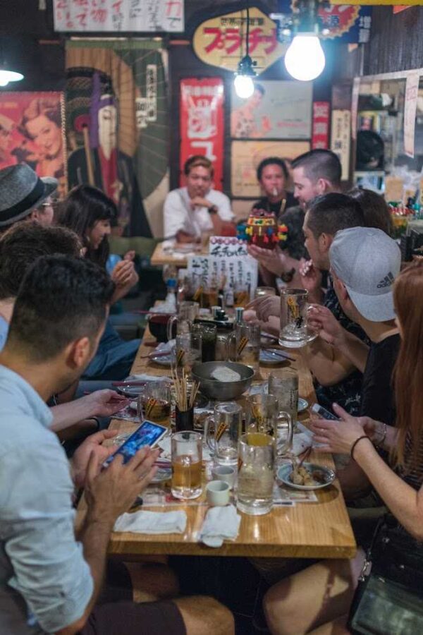 Okawari Travels guests sharing drinks around a long table in a Tokyo izakaya decorated with retro posters