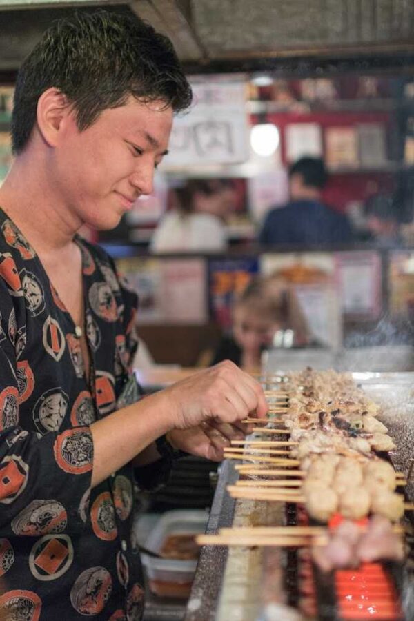 Cook in a patterned shirt turning yakitori skewers on a charcoal grill in a Tokyo izakaya