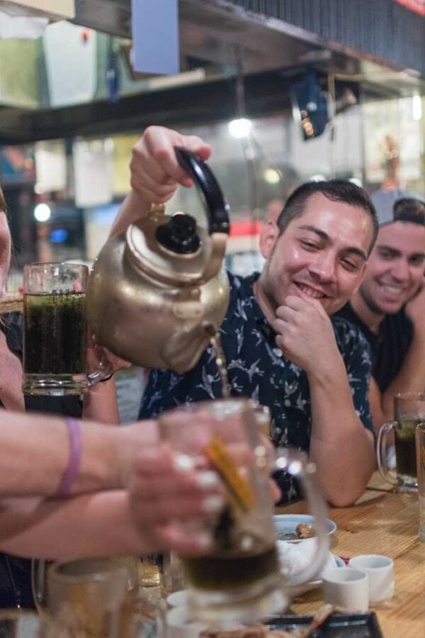 Guide pouring a drink from a metal kettle at a Tokyo izakaya table, with smiling guests behind