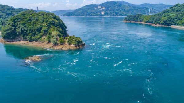 Wooded islands and swirling turquoise currents in a strait of the Seto Inland Sea, with a suspension bridge in the distance