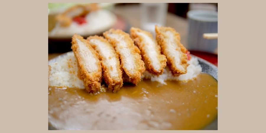 A close-up, side-angle shot of a plate of Japanese chicken katsu curry. Five thick, golden-brown breaded chicken cutlets are sliced and leaned against a mound of white rice. A generous portion of smooth, dark brown curry sauce pools at the bottom of the plate, with a small garnish of red pickled ginger visible on the side.