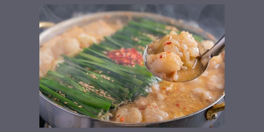 A steaming metal pot of Motsunabe, a famous Fukuoka hot pot. The dish features tender pieces of beef or pork offal (motsu) in a savory broth, topped with a generous layer of vibrant green Chinese chives, sprinkled with sesame seeds and red chili flakes. A ladle lifts a piece of the offal from the boiling soup.