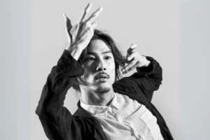 A black and white close up portrait of choreographer Kentaro Kujirai performing expressive hand movements and visceral facial expressions typical of butoh dance.