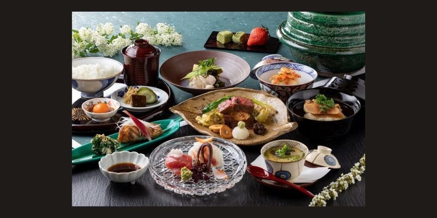 An elaborate and artistic spread of Kyoto Kaiseki cuisine on a dark table. The meal includes multiple small, decorative dishes such as sashimi in a crystal bowl, grilled Wagyu beef with seasonal vegetables, a bowl of clear dashi soup with tofu, and steamed rice. A red strawberry and matcha sweets are served as dessert in the background, all arranged with traditional Japanese aesthetic precision.
