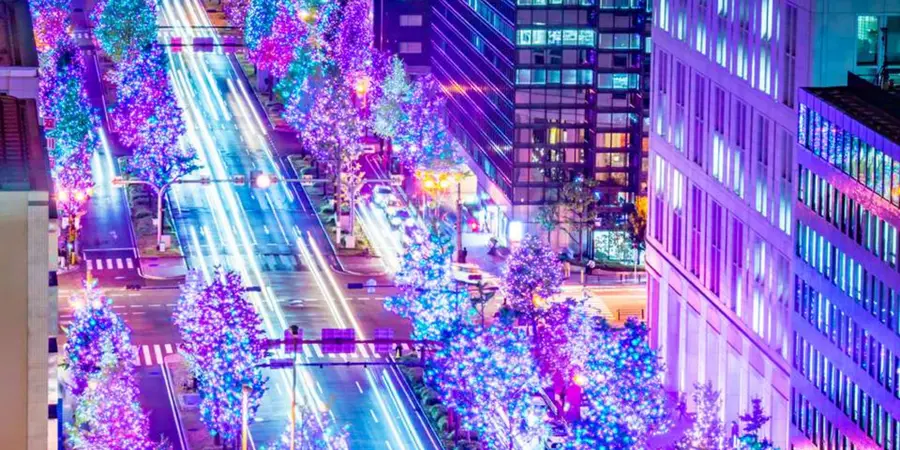 Osaka’s Midosuji Avenue lined with glowing trees; long-exposure light trails sweep between towers at night.