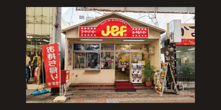 A street-level photograph of the "Jef" fast-food restaurant in Okinawa, Japan. The building has a prominent red and yellow sign that reads "Jef" in large letters with smaller Japanese text for "Goya Burger." The entrance is flanked by a red banner and a menu board, with several capsule toy (gachapon) machines sitting outside the glass front.