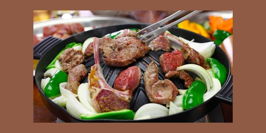 A close-up of a dome-shaped metal grill filled with Jingisukan, a Hokkaido specialty. Thick cuts of lamb and mutton are searing on the raised center of the grill, while a variety of fresh vegetables, including onions and green peppers, cook in the juices at the perimeter. Metal tongs are shown flipping a piece of meat.