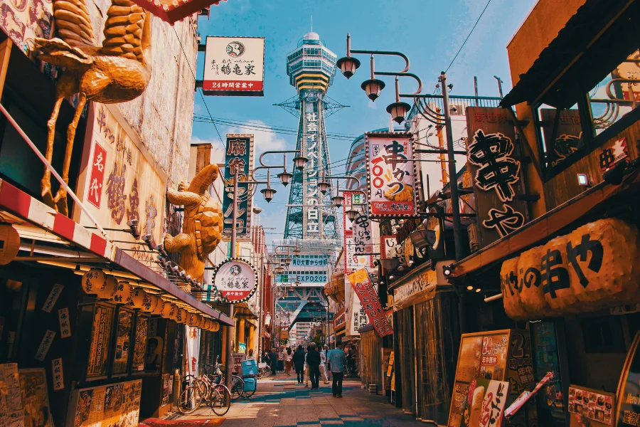 vibrant street in Osaka’s Shinsekai district, lined with colorful signs, lanterns, and traditional restaurants, with Tsutenkaku Tower standing tall in the background under a bright blue sky. The area is known for its retro charm, famous kushikatsu (deep-fried skewers), and nostalgic atmosphere.