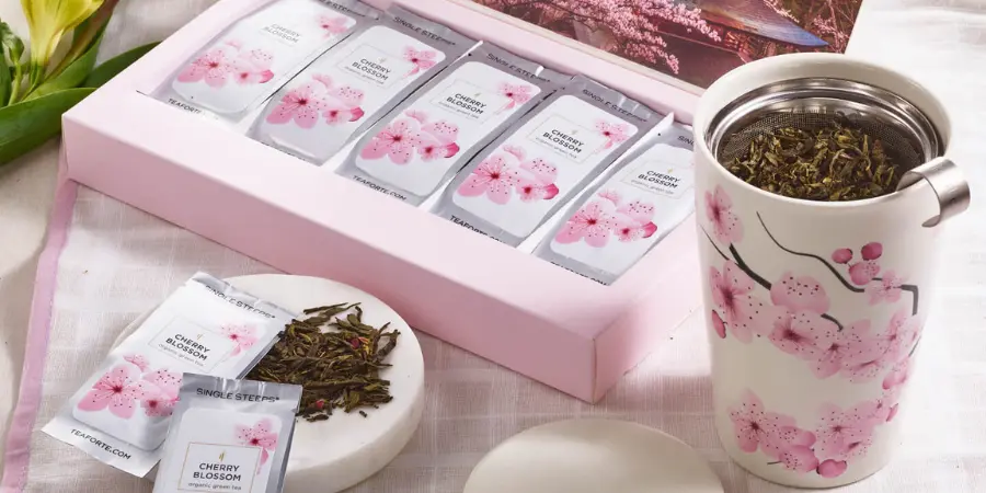 Tea packets are lined up in a box containing tea leaves infused with cherry blossoms. A pink and white cup is filled with sakura tea leaves.