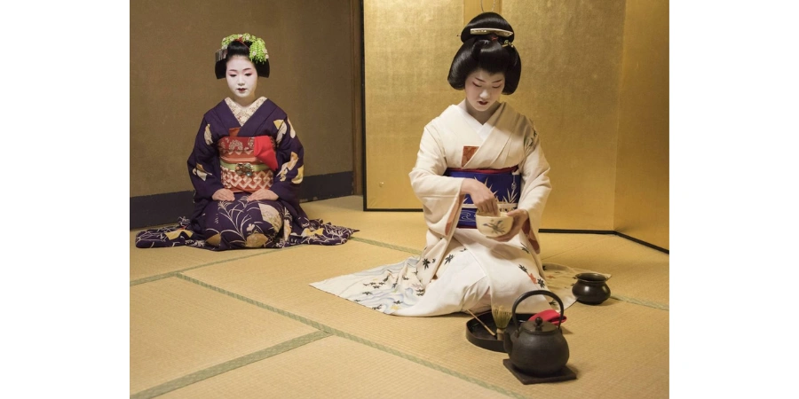 In a small room, two geishas are performing a traditional Japanese tea ceremony. One of them is mixing the green tea in a bowl.