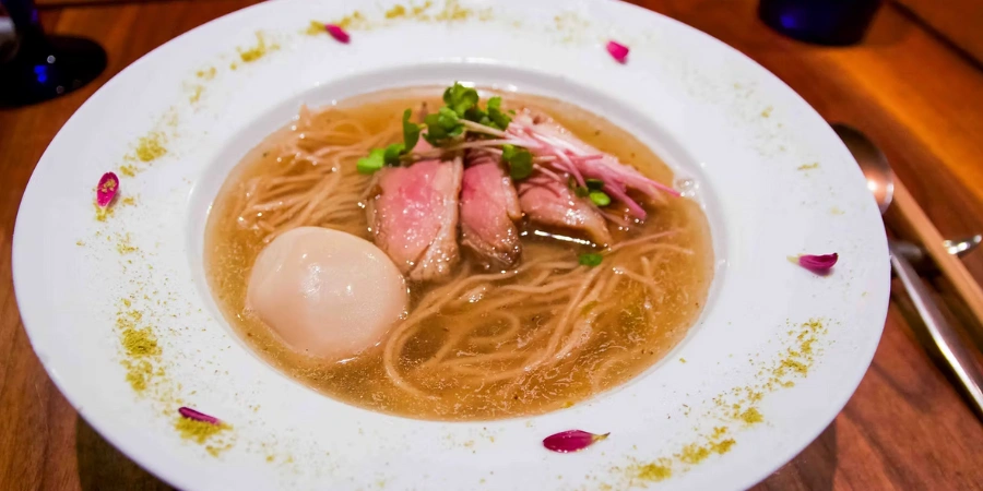 This is a bowl of ramen from Gion Duck Noodles. The ramen consists of a clear, golden broth, thin noodles, and slices of duck. It is topped off with a soft-boiled egg and microgreens with edible flowers on the rim of the plate.