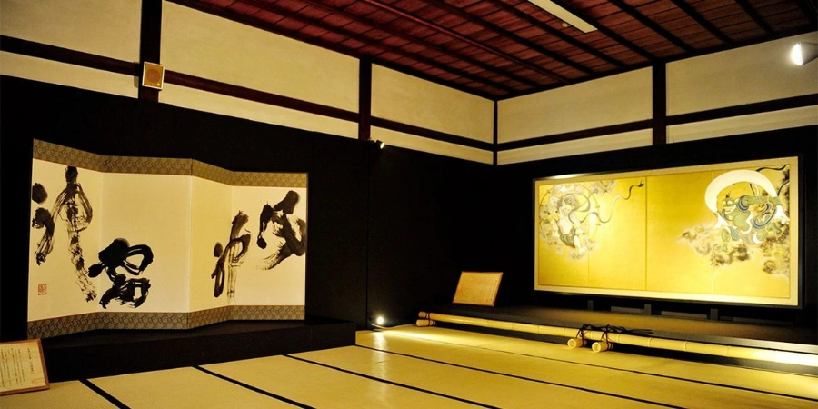 The room features traditional Japanese aesthetics, with tatami flooring and elegant fusuma (sliding doors) decorated with intricate artwork.The gold-leaf panel on the right depicts Raijin (the thunder god) and Fūjin (the wind god), two iconic deities from Japanese mythology, often associated with protection and nature’s power. The folding screen on the left displays bold calligraphy.