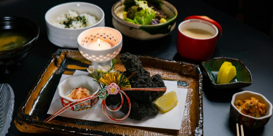 This image shows a traditional kaiseki meal. The black tempura-like dish in the foreground is identified as deep-fried fish or vegetables coated in a unique batter like bamboo charcoal. The candle and decorative ornaments add a refined, ceremonial touch.