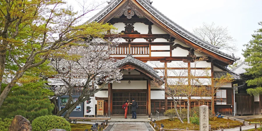The temple's main hall exhibits traditional Japanese architecture with a wooden framework, white plaster walls, and a gracefully sloping tiled roof adorned with ornate carvings. The entrance features a decorative gable with intricate woodwork. Surrounding the temple are lush gardens with manicured greenery, cherry blossoms, and maple trees, creating a serene and picturesque setting. In the foreground, two visitors are seen conversing near the entrance.