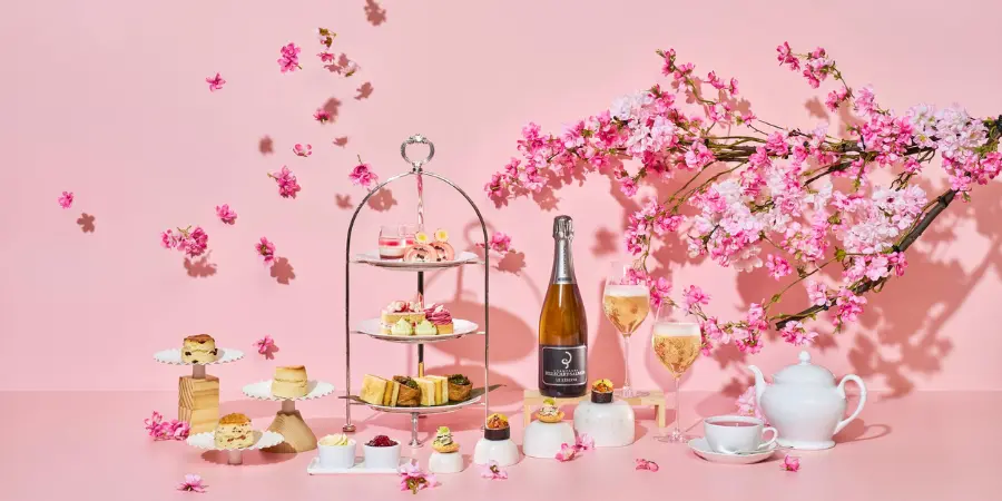 A full afternoon tea set is displayed behind a pink background with a small cherry blossom tree. The food consists of pastries and drinks made out of cherry blossoms.