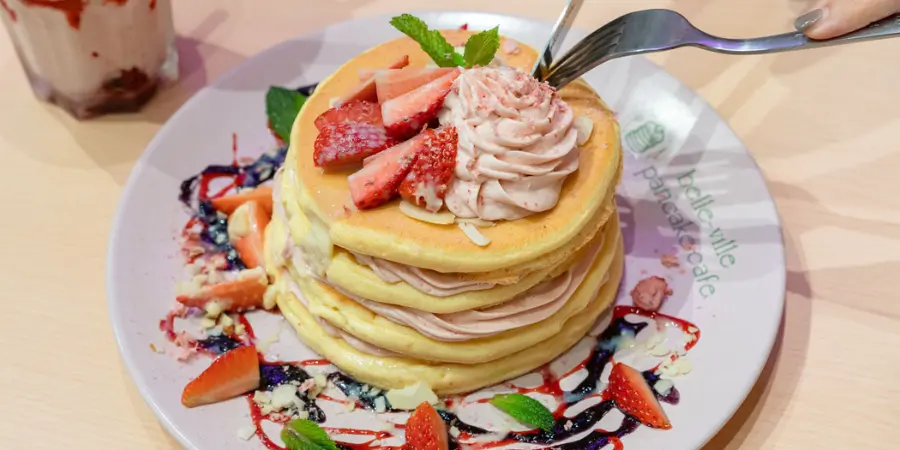 A plate of fluffy pancakes. There are slices of strawberry on the pancakes as well as around them. There is pink whipped cream on top.