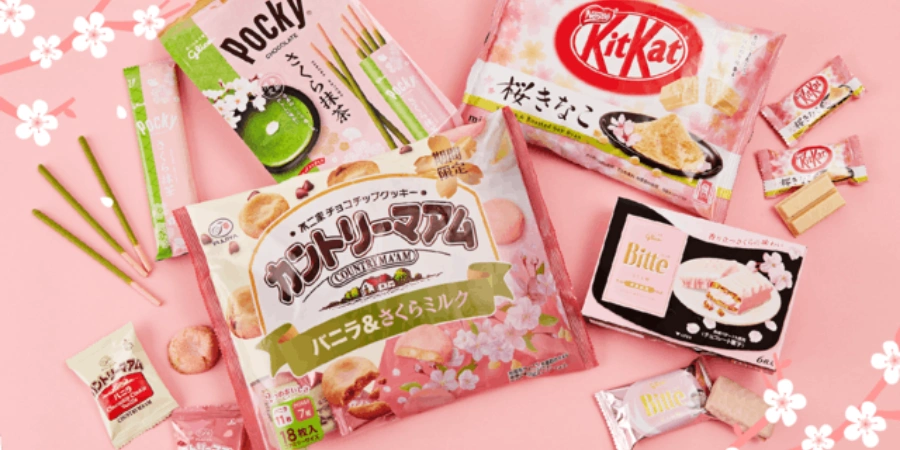 Different kinds of Japanese sakura-themed snacks are displayed on a pink surface. Some of these snacks include Pocky and KitKat.