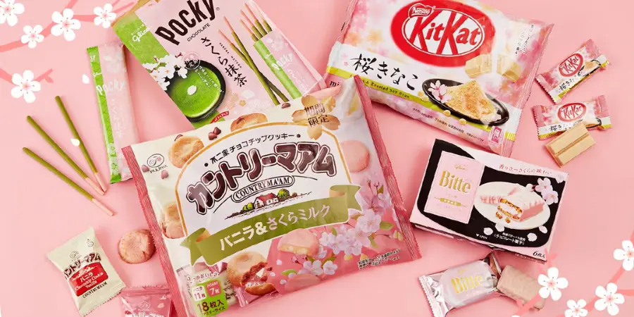 Different kinds of Japanese sakura-themed snacks are displayed on a pink surface. Some of these snacks include Pocky and KitKat.