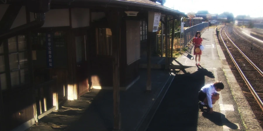 man tying his shoe lace at a regional japanese train station with a woman behind him on a sunny day in a rural Japanese trainstation