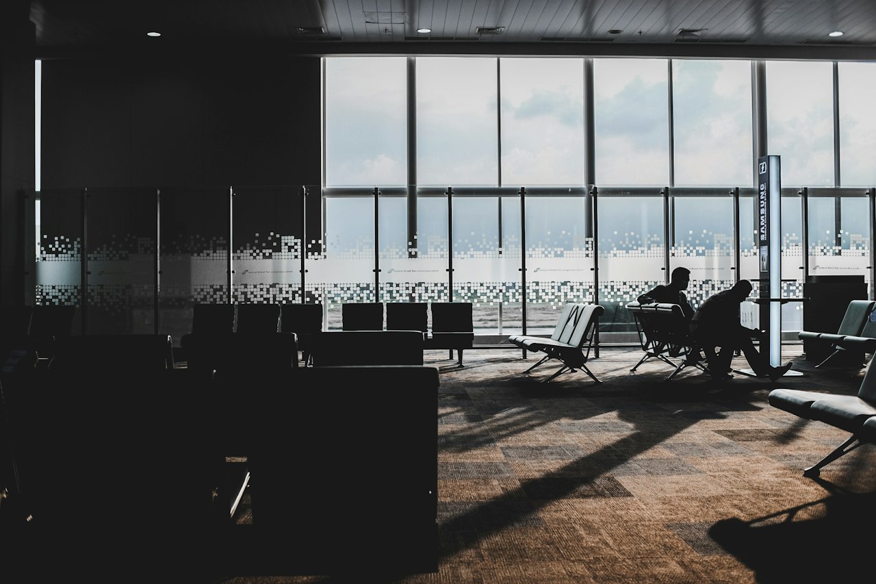 Travellers silhouetted on rows of seats beside tall windows at an airport departure gate