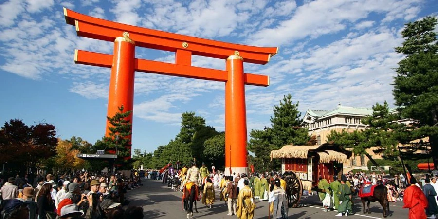 The great vermilion torii of Heian Shrine with the Jidai Matsuri procession passing beneath it, costumed participants and an ox-drawn cart on the road below