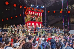 Bon Odori dancers performing around a traditional yagura stage under lanterns during a Japanese summer festival in August.