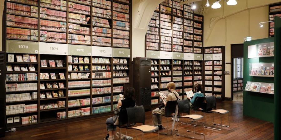Three visitors sit on chairs reading manga in front of towering, floor-to-ceiling shelves at the Kyoto International Manga Museum’s “Wall of Manga.”