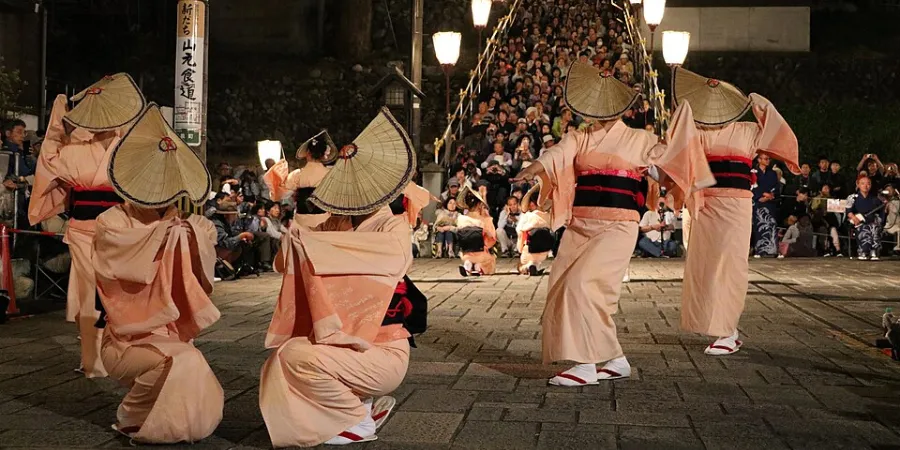 Traditional Japanese dancers wearing straw hats and pastel kimono perform at the Owara Kaze no Bon festival in Toyama, Japan, surrounded by a large nighttime crowd.