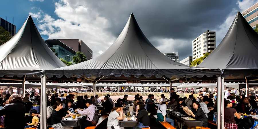 Busy outdoor food festival in Sapporo, Japan, with visitors dining beneath large pointed gray canopy tents, surrounded by modern city buildings and dramatic cloudy skies.