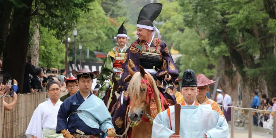 A mounted samurai in ornate traditional Japanese armor leads a ceremonial horse procession through the forested grounds of Tsurugaoka Hachimangu Shrine in Kamakura, Japan, accompanied by participants in historical costumes and watched by spectators.