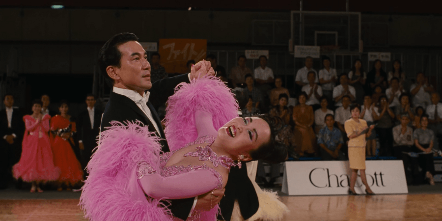 Shohei Sugiyama (Kōji Yakusho) and his dance partner, Toyoko Takahashi, performing a graceful but hesitant social dance step during a crowded local ballroom competition.