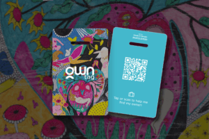 A front and back view of the OWN Tag smart luggage tag featuring a vibrant artistic design on the front and a QR code with NFC technology on the back. The tag allows a finder to tap or scan to help find the owner without exposing private contact details.