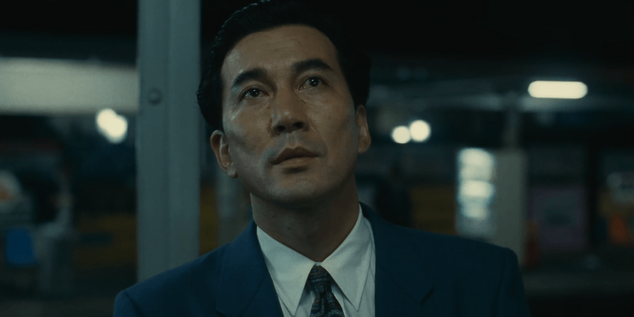 A mid-range shot of Shohei Sugiyama (Kōji Yakusho) standing on a dimly lit street, looking up with subtle, yearning eyes at the brightly lit dance school entrance sign.