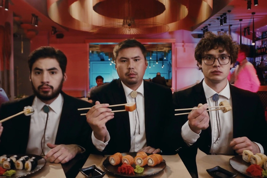 Three men in suits and bolo ties holding sushi rolls with chopsticks at a vibrant Japanese restaurant in London. This highlights the exciting journey for food lovers exploring diverse flavors from sushi bars to authentic ramen shops across the city.