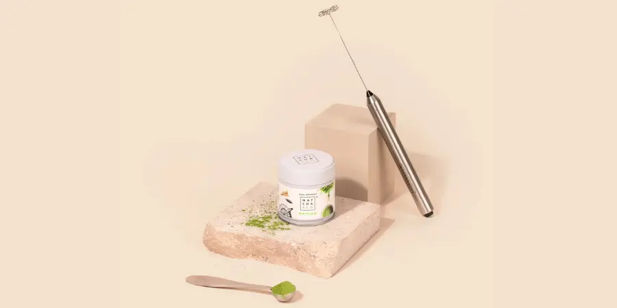 A container of Matcha & Co. powder and a measuring spoon resting on a beige stone block, next to a silver handheld electric milk frother.