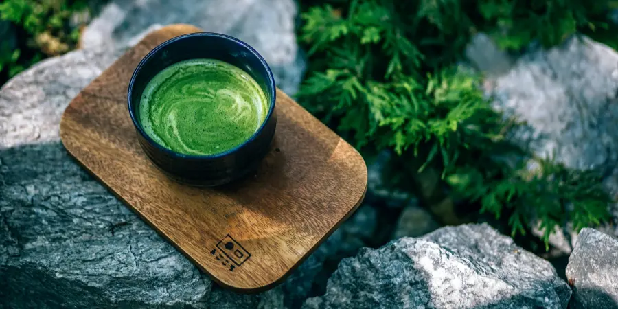 A dark ceramic bowl of hot matcha tea with foam art, resting on a small wooden tray placed on large gray rocks outdoors.