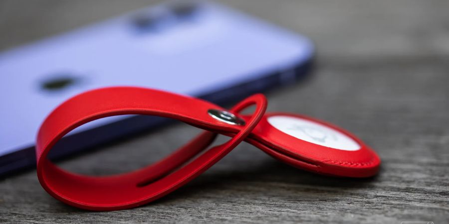 lose-up of a white Apple AirTag housed within a red leather loop, resting on a wooden surface with a purple smartphone visible in the soft background.