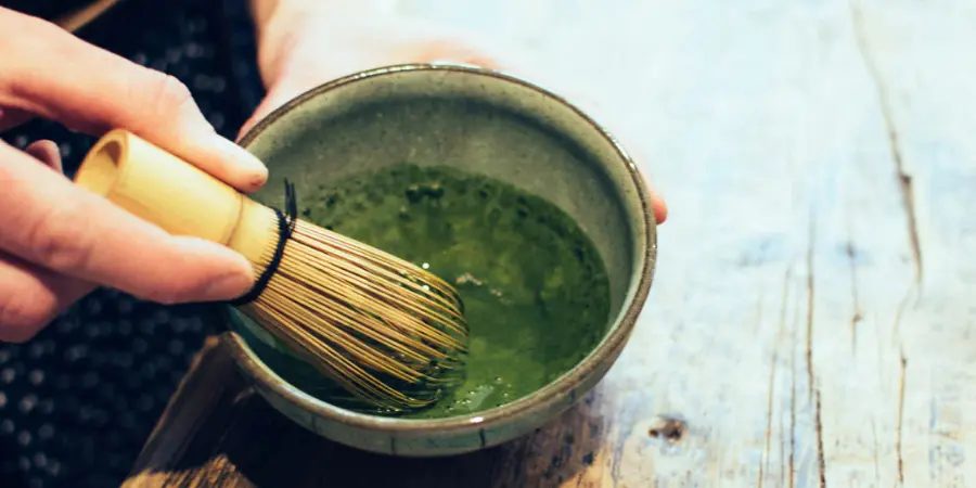 Close-up of a bamboo whisk (*chasen*) being used to rapidly whisk matcha tea and water into a smooth, frothy consistency in a ceramic bowl.