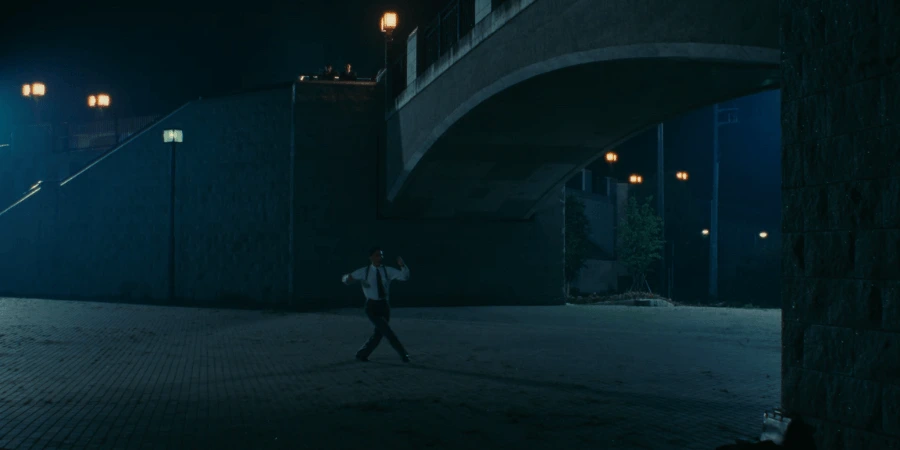 Shohei Sugiyama practicing a hesitant social dance step under a bridge, unaware that the hired Private Investigator is secretly watching his movements from a hidden position.